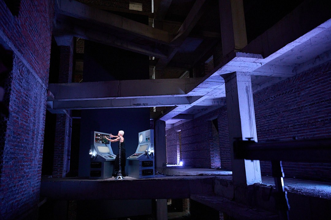 A distant wide view across the unfinished concrete building towards M07 performing under violet light