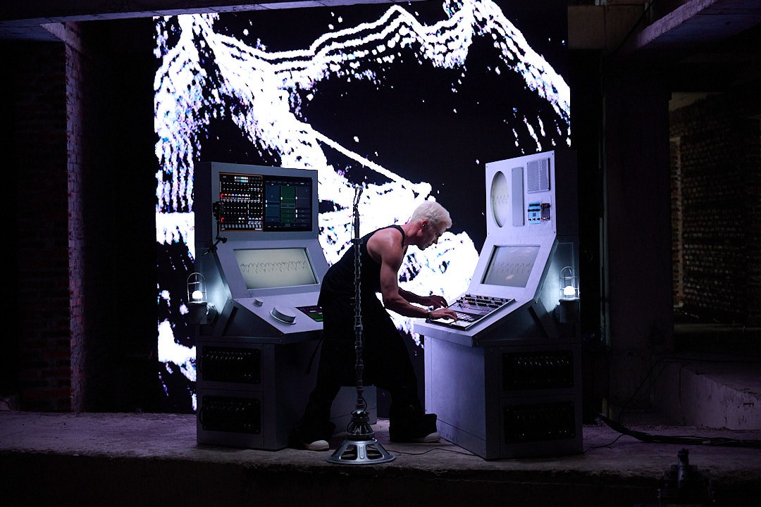 M07 in movement between the custom consoles with a large monochrome face on the screen behind the performance