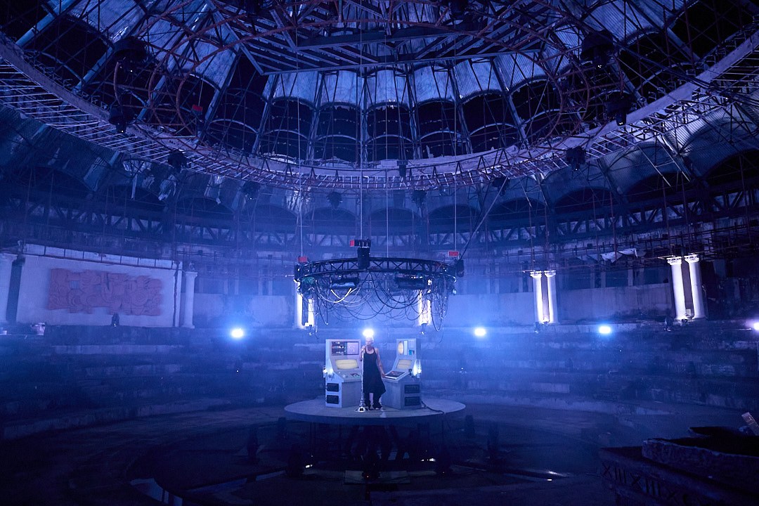 Wide view of the abandoned circular circus, with M07 and the instrument platform under blue light beneath the surrounding tiers